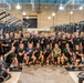 A large group of approximately 40-50 individuals, predominantly wearing black military-style athletic shirts with 'ARMY' branding, pose for a group photo inside a well-equipped military fitness facility. Many individuals are making 'number one' finger gestures toward the camera, suggesting a celebratory or team-unity moment. The facility features rows of black and yellow Rogue fitness equipment including power racks, cable machines, and assault bikes.