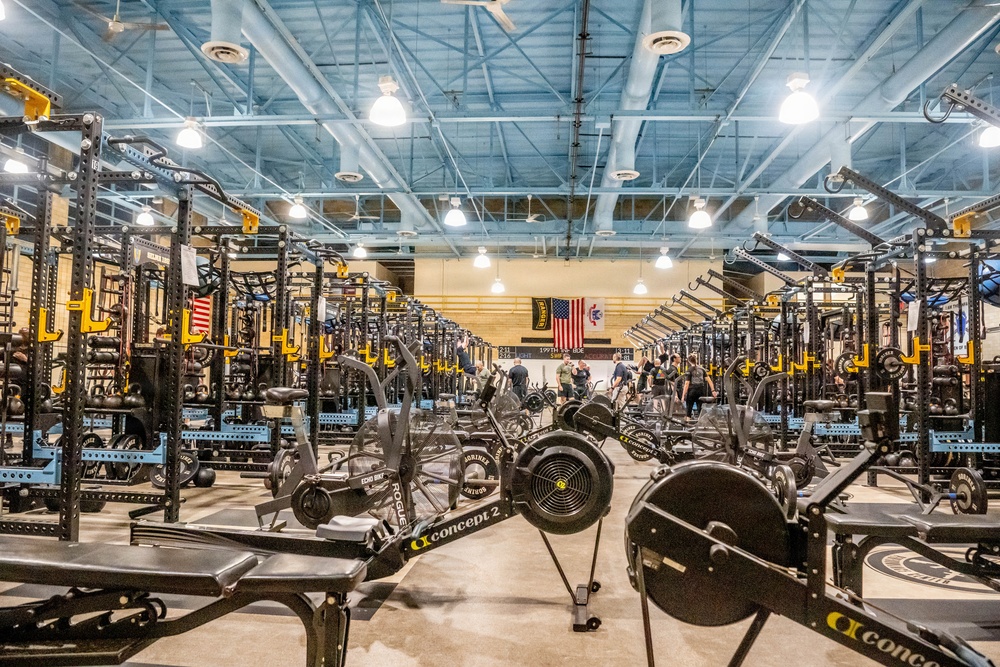 A large, well-equipped military or institutional weight room featuring rows of Rogue power racks with yellow safety arms, Concept2 rowing machines, and air bikes stretching deep into the facility. An American flag and what appears to be a unit or organizational banner are displayed prominently on the far wall. Several individuals are visible in the background actively using the equipment.