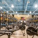 A large, well-equipped military or institutional weight room featuring rows of Rogue power racks with yellow safety arms, Concept2 rowing machines, and air bikes stretching deep into the facility. An American flag and what appears to be a unit or organizational banner are displayed prominently on the far wall. Several individuals are visible in the background actively using the equipment.