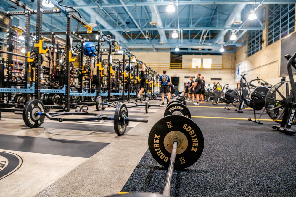 A large, professional-grade athletic weight room featuring rows of power racks with black and yellow color scheme. The facility is equipped with Sorinex-branded barbells and weight plates in the foreground, with multiple athletes visible in the background. The space appears to be a collegiate or professional sports training facility with high ceilings, industrial lighting, and specialized rubber flooring.