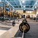 A large, professional-grade athletic weight room featuring rows of power racks with black and yellow color scheme. The facility is equipped with Sorinex-branded barbells and weight plates in the foreground, with multiple athletes visible in the background. The space appears to be a collegiate or professional sports training facility with high ceilings, industrial lighting, and specialized rubber flooring.