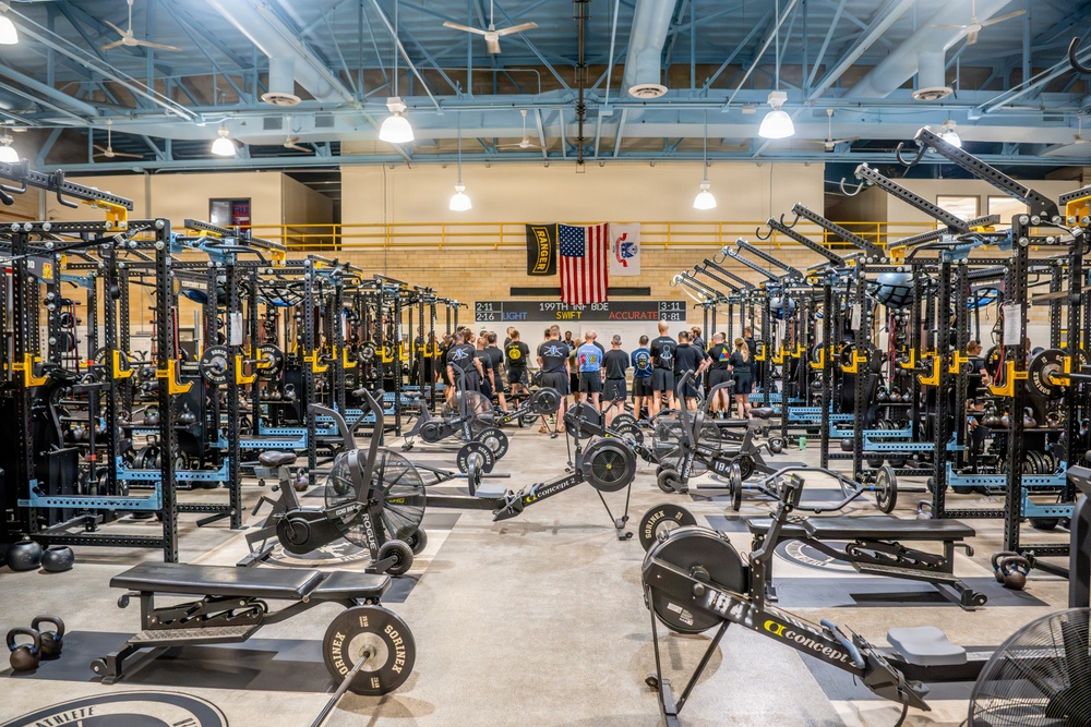 A large, well-equipped military fitness facility, likely belonging to the 199th Infantry Brigade based on visible signage. The gym features rows of power racks, rowing machines, air bikes, and free weights arranged in an organized layout. A group of personnel dressed in dark PT (physical training) uniforms are gathered at the far end of the facility near an American flag and unit insignia.