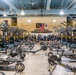 A large, well-equipped military fitness facility, likely belonging to the 199th Infantry Brigade based on visible signage. The gym features rows of power racks, rowing machines, air bikes, and free weights arranged in an organized layout. A group of personnel dressed in dark PT (physical training) uniforms are gathered at the far end of the facility near an American flag and unit insignia.