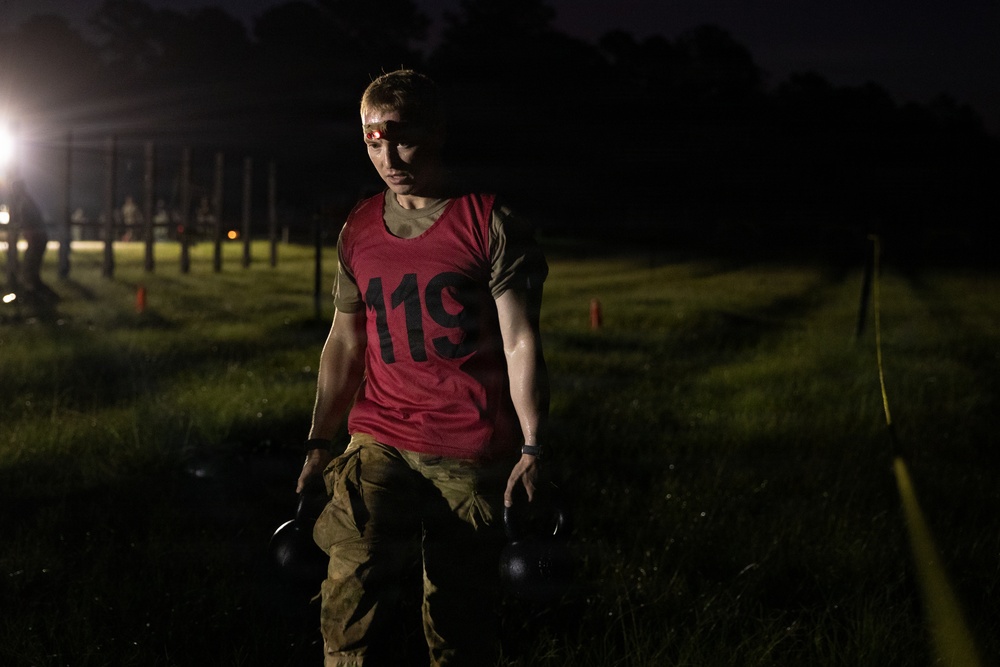 A person wearing a red numbered bib (119) and military-style camouflage pants carries two kettlebells during what appears to be a nighttime fitness or military assessment event. They have a red headlamp on their forehead and appear physically exerted, with sweat visible on their face and clothing. The setting is an outdoor grassy field illuminated by artificial lighting in low-light conditions.