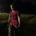 A person wearing a red numbered bib (119) and military-style camouflage pants carries two kettlebells during what appears to be a nighttime fitness or military assessment event. They have a red headlamp on their forehead and appear physically exerted, with sweat visible on their face and clothing. The setting is an outdoor grassy field illuminated by artificial lighting in low-light conditions.