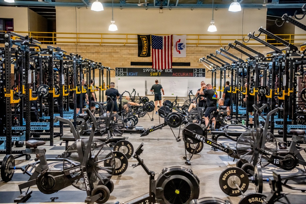 A large military fitness facility, identified as belonging to the 199th Infantry Brigade, is equipped with extensive strength and conditioning equipment. The gym features rows of Rogue power racks, Concept2 rowing machines, Echo Bikes, and Sorinex weight plates. Several individuals dressed in black PT (physical training) uniforms are actively working out or moving through the space.