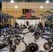 A large military fitness facility, identified as belonging to the 199th Infantry Brigade, is equipped with extensive strength and conditioning equipment. The gym features rows of Rogue power racks, Concept2 rowing machines, Echo Bikes, and Sorinex weight plates. Several individuals dressed in black PT (physical training) uniforms are actively working out or moving through the space.
