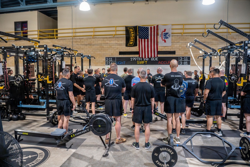A large group of military personnel in matching black PT (physical training) uniforms are gathered inside a well-equipped military fitness facility. The setting appears to be a briefing or formation before a workout session, with the group facing a speaker at the center. The gym is associated with the 199th Infantry Brigade, as indicated by signage and flags on the wall.