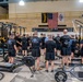 A large group of military personnel in matching black PT (physical training) uniforms are gathered inside a well-equipped military fitness facility. The setting appears to be a briefing or formation before a workout session, with the group facing a speaker at the center. The gym is associated with the 199th Infantry Brigade, as indicated by signage and flags on the wall.