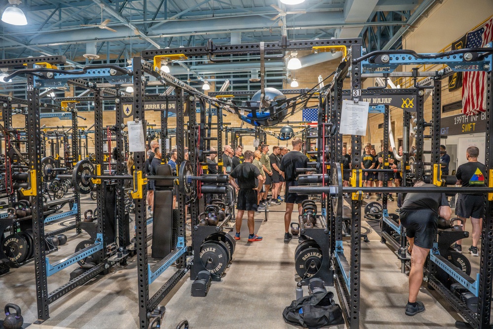 A large military fitness facility filled with rows of Sorinex power racks and weightlifting equipment. A group of personnel, predominantly wearing black PT (physical training) uniforms, are gathered in the center aisle, likely receiving a briefing or orientation. An American flag and unit battle streamers, including a reference to '199th INF BN' and 'AN NAJAF,' are visible on the far wall.