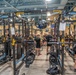 A large military fitness facility filled with rows of Sorinex power racks and weightlifting equipment. A group of personnel, predominantly wearing black PT (physical training) uniforms, are gathered in the center aisle, likely receiving a briefing or orientation. An American flag and unit battle streamers, including a reference to '199th INF BN' and 'AN NAJAF,' are visible on the far wall.