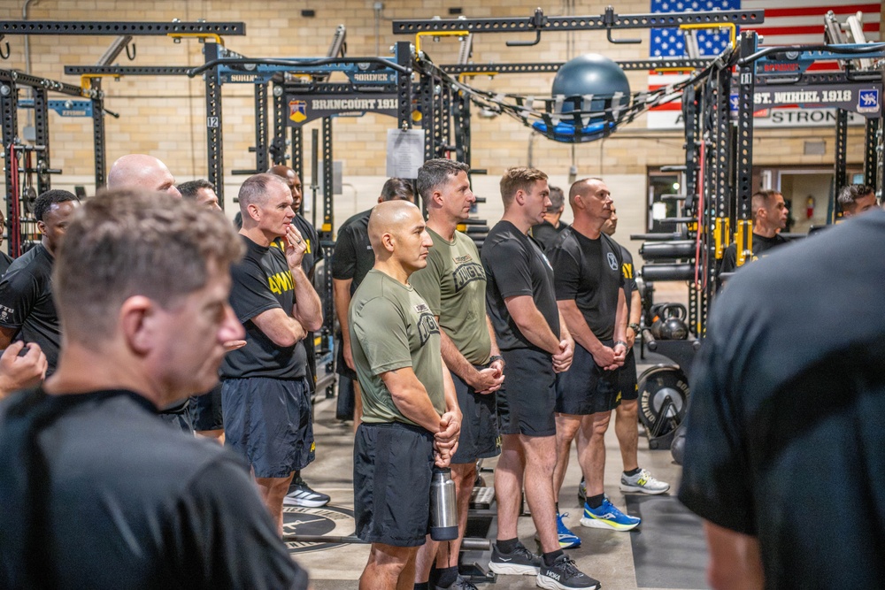 A group of military personnel, predominantly wearing black and olive/green athletic attire with 'Army' branding, are gathered in a well-equipped military fitness facility. They appear to be standing at attention or listening to a briefing, arranged in rows facing an unseen speaker. The facility features heavy-duty power racks, weight equipment, and military battle streamers/signage on the walls including references to 'BRANCOURT 1918' and 'ST. MIHIEL 1918'.