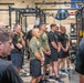 A group of military personnel, predominantly wearing black and olive/green athletic attire with 'Army' branding, are gathered in a well-equipped military fitness facility. They appear to be standing at attention or listening to a briefing, arranged in rows facing an unseen speaker. The facility features heavy-duty power racks, weight equipment, and military battle streamers/signage on the walls including references to 'BRANCOURT 1918' and 'ST. MIHIEL 1918'.