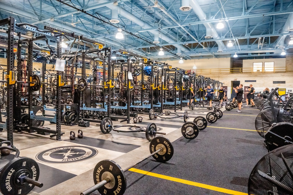 A large, professional-grade strength and conditioning facility featuring rows of black power racks with yellow accents, branded 'Sorinex' equipment throughout. The facility appears to be a collegiate or military athletic training center, with numerous barbells, weight plates, and kettlebells arranged on the floor. Several individuals are visible in the background engaged in training or coaching activities.