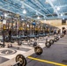 A large, professional-grade strength and conditioning facility featuring rows of black power racks with yellow accents, branded 'Sorinex' equipment throughout. The facility appears to be a collegiate or military athletic training center, with numerous barbells, weight plates, and kettlebells arranged on the floor. Several individuals are visible in the background engaged in training or coaching activities.