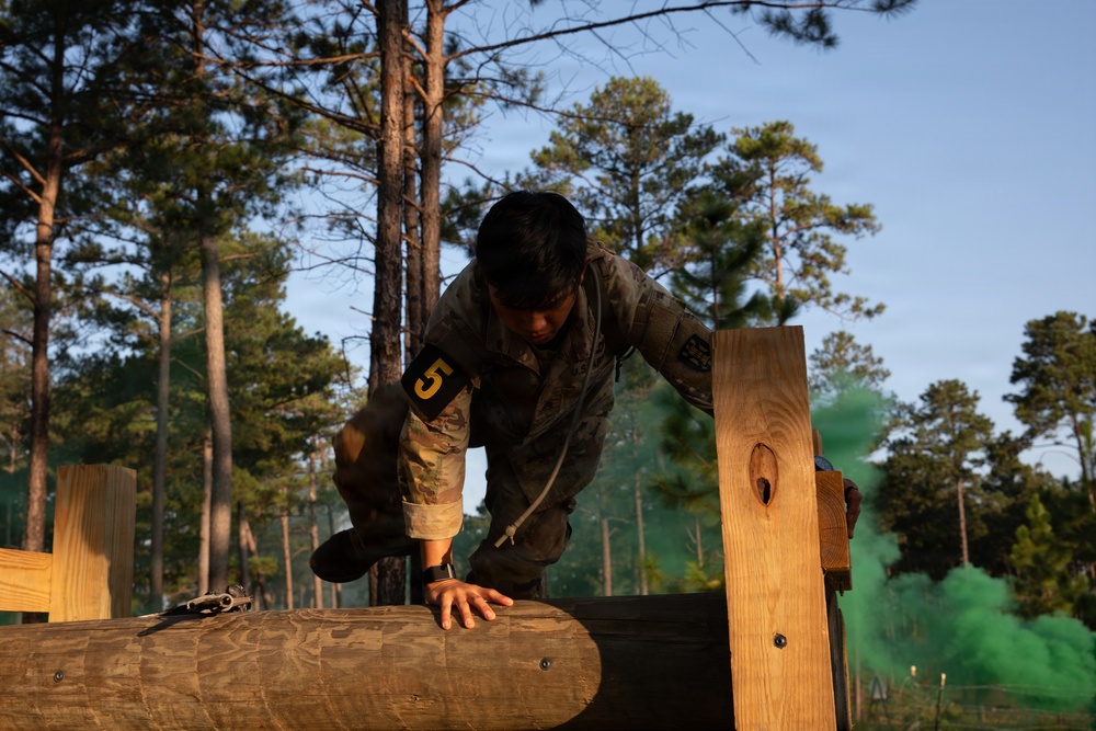 A soldier wearing camouflage uniform and numbered bib '5' is navigating a military obstacle course, climbing over a large log barrier. Green smoke can be seen in the background, suggesting a simulated combat or training exercise environment. The setting appears to be a wooded military training area during early morning or late afternoon light.