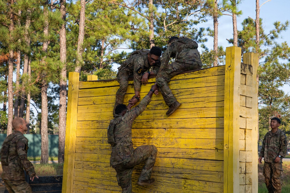 Military personnel in camouflage uniforms are navigating a tall yellow wooden wall obstacle as part of what appears to be a military training exercise or obstacle course. Soldiers at the top of the wall are pulling up a teammate who is climbing from below, demonstrating teamwork and physical fitness training. Additional soldiers observe from the sides of the obstacle.