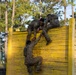 Military personnel in camouflage uniforms are navigating a tall yellow wooden wall obstacle as part of what appears to be a military training exercise or obstacle course. Soldiers at the top of the wall are pulling up a teammate who is climbing from below, demonstrating teamwork and physical fitness training. Additional soldiers observe from the sides of the obstacle.