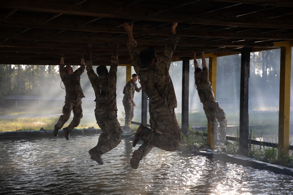 Military personnel in camouflage uniforms are traversing an obstacle course, hanging and moving hand-over-hand across the underside of a wooden overhead structure above a water pit. The scene is backlit with atmospheric haze or mist, creating dramatic lighting. One instructor or observer stands upright in the background monitoring the activity.