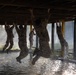 Military personnel in camouflage uniforms are traversing an obstacle course, hanging and moving hand-over-hand across the underside of a wooden overhead structure above a water pit. The scene is backlit with atmospheric haze or mist, creating dramatic lighting. One instructor or observer stands upright in the background monitoring the activity.