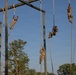 Multiple military personnel in camouflage uniforms are climbing vertical ropes on a large wooden obstacle course structure. The training apparatus consists of tall wooden poles with horizontal crossbeams from which several thick ropes hang. The scene appears to be an outdoor military physical training exercise taking place during daylight hours.