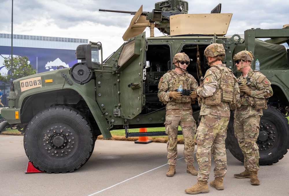 Three U.S. Army soldiers in MultiCam (OCP) uniforms and combat gear are gathered near a military tactical vehicle, appearing to be at a public display or demonstration event. The vehicle, marked 'D0063', appears to be a Joint Light Tactical Vehicle (JLTV) equipped with a remote weapon station on top. The setting appears to be an outdoor venue with civilian infrastructure visible in the background.