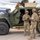 Three U.S. Army soldiers in MultiCam (OCP) uniforms and combat gear are gathered near a military tactical vehicle, appearing to be at a public display or demonstration event. The vehicle, marked 'D0063', appears to be a Joint Light Tactical Vehicle (JLTV) equipped with a remote weapon station on top. The setting appears to be an outdoor venue with civilian infrastructure visible in the background.