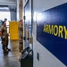 The image shows military personnel in camouflage uniforms gathered in what appears to be a military facility near an armory. A prominent blue door or wall panel with yellow lettering reading 'ARMORY' is visible in the foreground. Several soldiers are standing and conversing in the background near a large open bay door.
