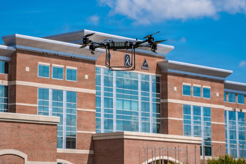 A quadcopter drone is captured mid-flight in front of a modern multi-story brick building with large glass windows. The drone appears to be a professional or research-grade model with four rotors and a rectangular frame. The building in the background features institutional signage and architectural emblems, suggesting a government, military, or academic facility.