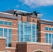 A quadcopter drone is captured mid-flight in front of a modern multi-story brick building with large glass windows. The drone appears to be a professional or research-grade model with four rotors and a rectangular frame. The building in the background features institutional signage and architectural emblems, suggesting a government, military, or academic facility.