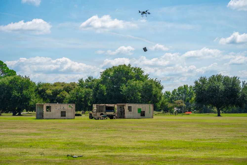 A multi-rotor drone is captured mid-flight over an open grassy field, appearing to drop or release a package. Below, two modified shipping containers with green markings and a military-style vehicle (likely a HMMWV/Humvee) are arranged in a simulated compound or training environment. The setting suggests a drone delivery or military logistics training exercise.