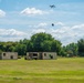 A multi-rotor drone is captured mid-flight over an open grassy field, appearing to drop or release a package. Below, two modified shipping containers with green markings and a military-style vehicle (likely a HMMWV/Humvee) are arranged in a simulated compound or training environment. The setting suggests a drone delivery or military logistics training exercise.