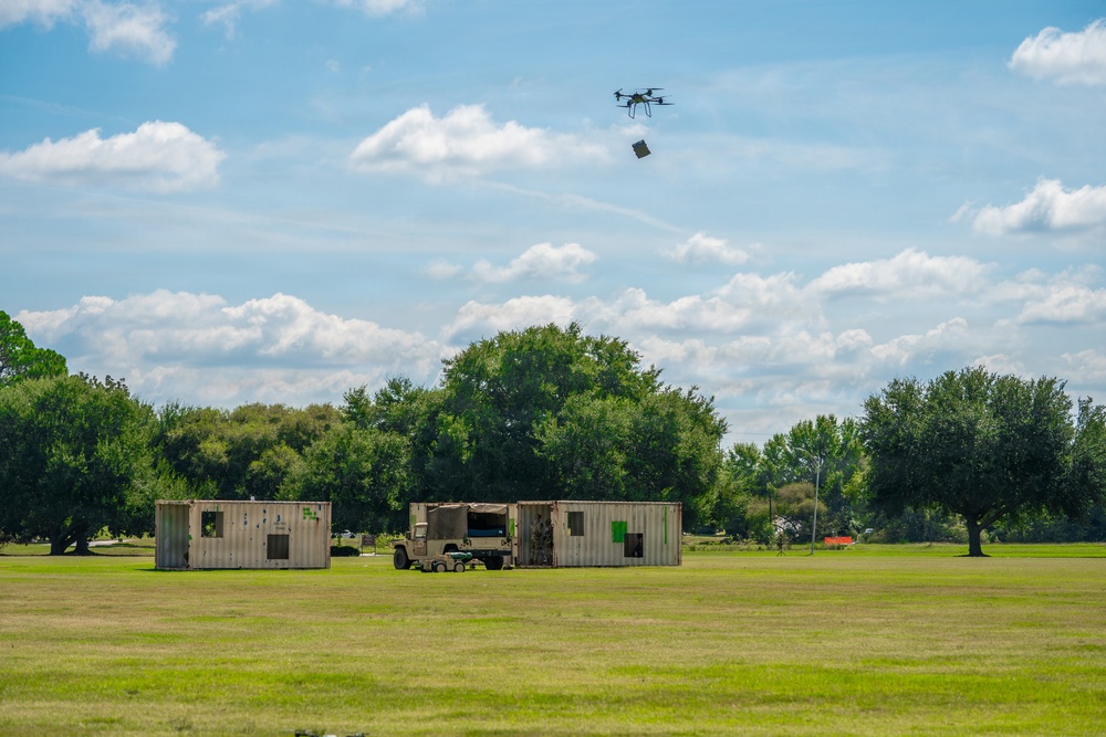 A multi-rotor drone is captured mid-flight over an open grassy field, appearing to drop or deliver a small package. Below, two shipping containers and a military-style vehicle (likely a Humvee) are positioned on the field, suggesting a logistics or resupply demonstration. The setting appears to be a military or government testing/training exercise.