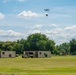 A multi-rotor drone is captured mid-flight over an open grassy field, appearing to drop or deliver a small package. Below, two shipping containers and a military-style vehicle (likely a Humvee) are positioned on the field, suggesting a logistics or resupply demonstration. The setting appears to be a military or government testing/training exercise.