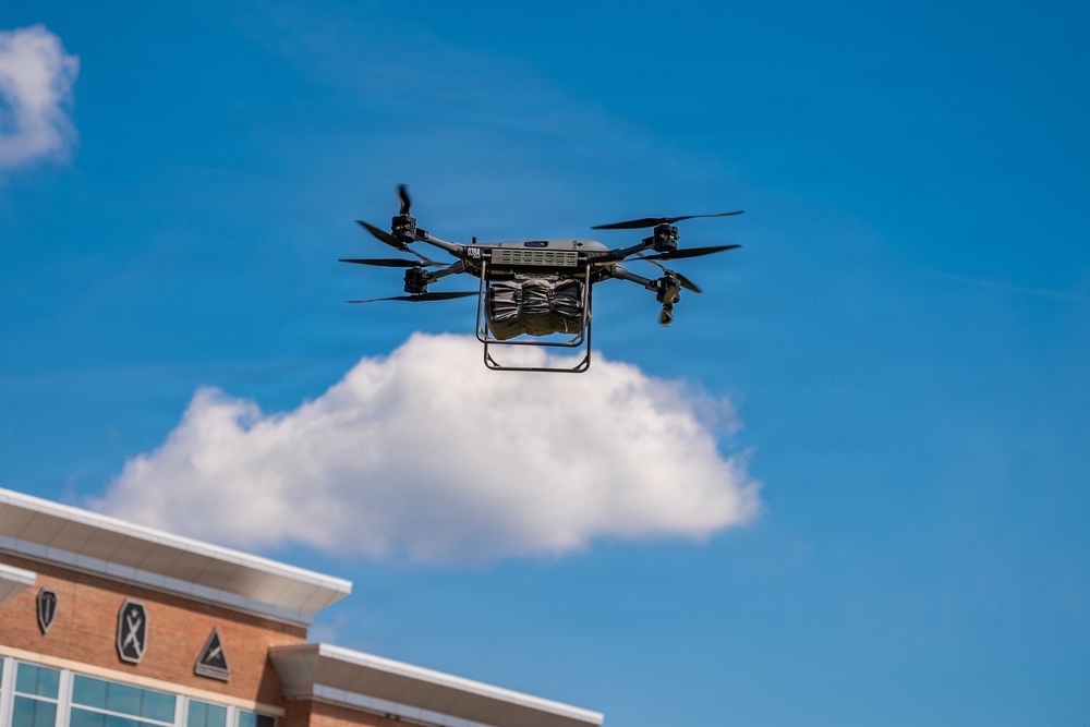A commercial delivery drone is captured mid-flight against a bright blue sky with scattered white clouds. The quadcopter-style drone is carrying a cargo package suspended beneath its frame, flying above what appears to be a commercial or institutional building. The image is taken from a low angle looking upward, providing a clear view of the drone's rotor configuration and payload.