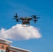 A commercial delivery drone is captured mid-flight against a bright blue sky with scattered white clouds. The quadcopter-style drone is carrying a cargo package suspended beneath its frame, flying above what appears to be a commercial or institutional building. The image is taken from a low angle looking upward, providing a clear view of the drone's rotor configuration and payload.
