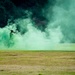 The image shows what appears to be a military or tactical training exercise taking place on an open grass field. Two figures in military-style gear are partially obscured by a large cloud of bright green smoke, likely from a smoke grenade used for concealment or signaling. The scene is set against a backdrop of dark trees.