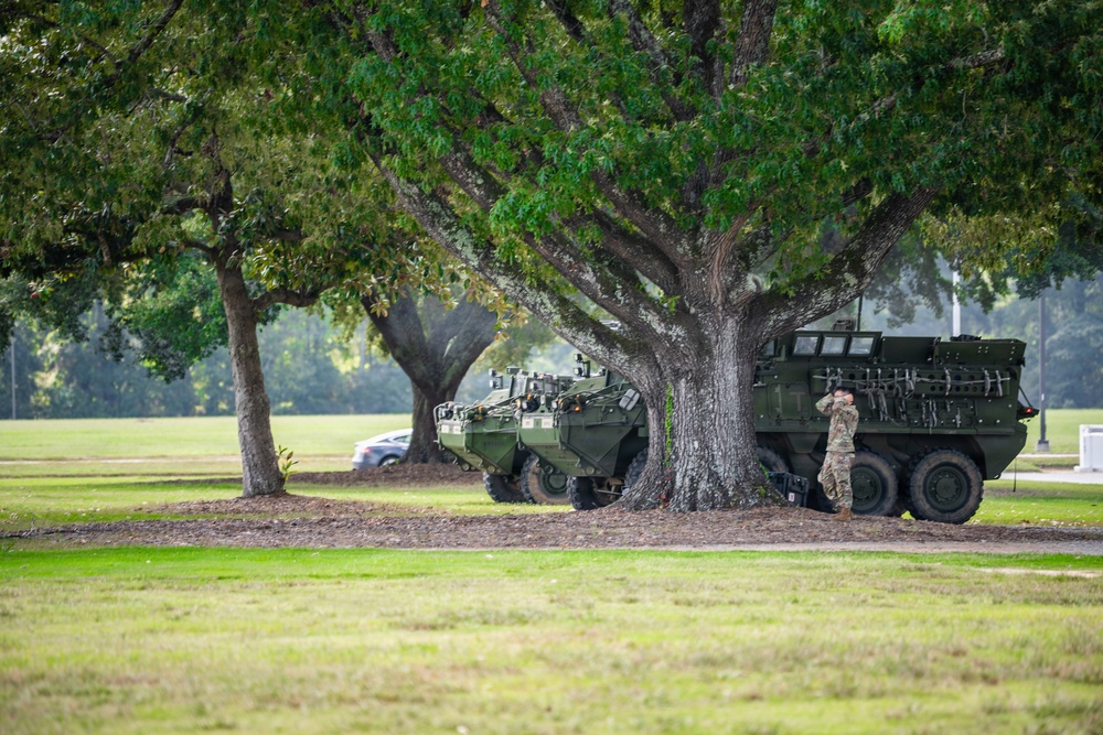 The image shows two military armored vehicles, consistent with Stryker-type wheeled armored personnel carriers, parked beneath large oak trees on a grassy field. At least one soldier in camouflage uniform is visible standing near the vehicles. The setting appears to be a military installation or park-like grounds.
