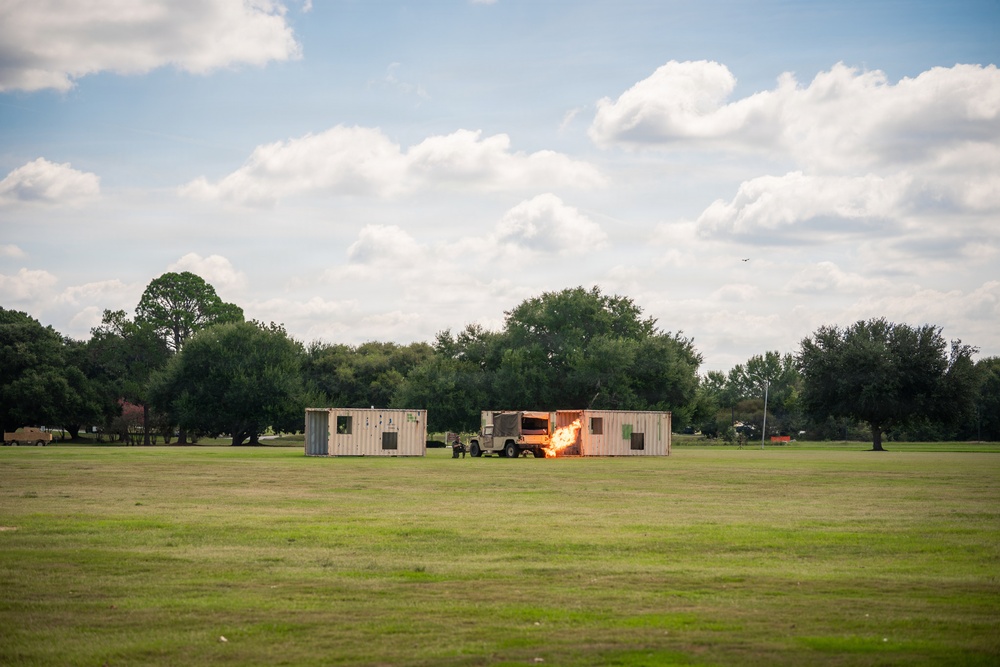 A military training exercise or demonstration is taking place on a large open grass field. Two shipping containers are positioned on the field with a military vehicle (appears to be a Humvee) between them, with a visible explosion or pyrotechnic blast occurring at the junction point. The scene is set against a backdrop of trees under a partly cloudy sky.