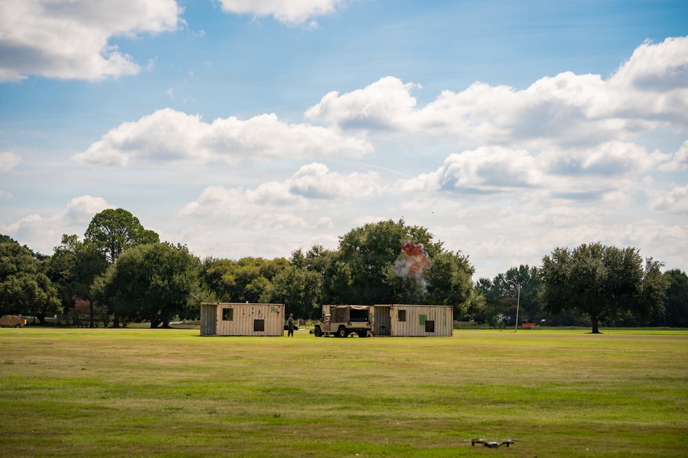 A military training exercise is taking place on an open grassy field, featuring two tan shipping containers arranged as a mock compound with a military HMMWV (Humvee) positioned between them. An explosion or pyrotechnic detonation is visible behind the right container, producing orange and white smoke. A small drone is visible in the lower right foreground, likely used for observation or documentation of the exercise.