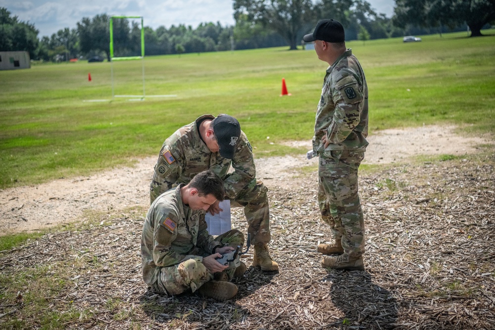 Three U.S. Army soldiers in OCP (Operational Camouflage Pattern) uniforms are gathered on a grassy field, with two crouching over what appears to be a drone controller or electronic device while a third stands observing. The setting appears to be a training exercise on a military installation, evidenced by orange traffic cones and a goal post structure in the background. The soldiers are wearing American flag patches and unit patches consistent with U.S. Army affiliation.
