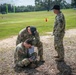 Three U.S. Army soldiers in OCP (Operational Camouflage Pattern) uniforms are gathered on a grassy field, with two crouching over what appears to be a drone controller or electronic device while a third stands observing. The setting appears to be a training exercise on a military installation, evidenced by orange traffic cones and a goal post structure in the background. The soldiers are wearing American flag patches and unit patches consistent with U.S. Army affiliation.