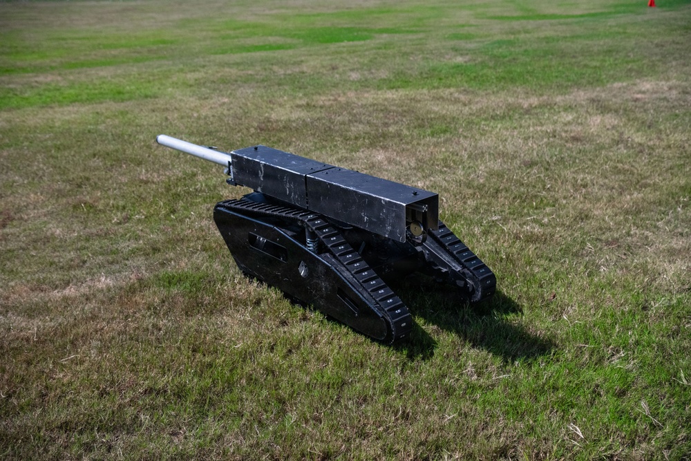 The image shows a small black unmanned ground vehicle (UGV) or robotic platform mounted on rubber tank-style tracks, equipped with a cylindrical barrel or tube on top. The device appears to be a miniature robotic tank, likely a prototype or demonstration unit, positioned on a grassy field. Its construction appears to be largely custom-built or fabricated from metal components.