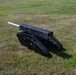 The image shows a small black unmanned ground vehicle (UGV) or robotic platform mounted on rubber tank-style tracks, equipped with a cylindrical barrel or tube on top. The device appears to be a miniature robotic tank, likely a prototype or demonstration unit, positioned on a grassy field. Its construction appears to be largely custom-built or fabricated from metal components.