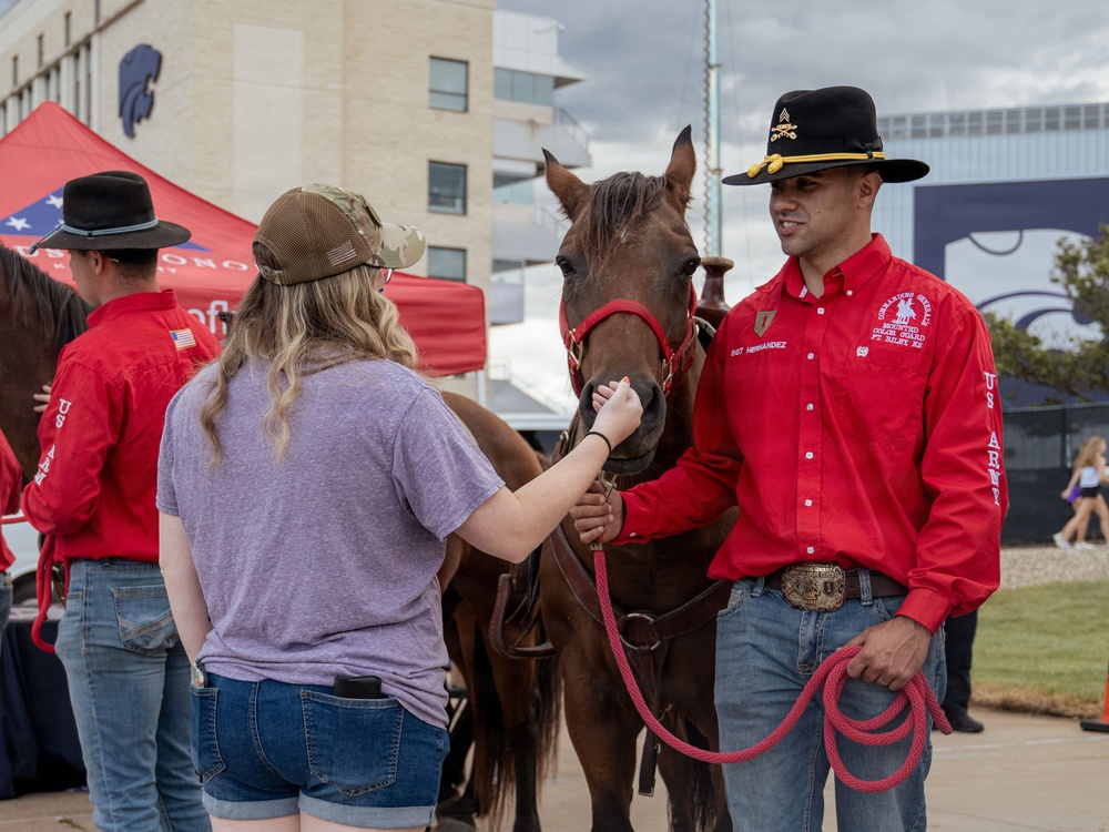 A U.S. Army soldier identified as SGT Hernandez from the Commanding General's Mounted Color Guard at Fort Riley, Kansas, is allowing a young woman to pet a dark brown horse at what appears to be a public outreach event. The soldier is wearing a distinctive red western shirt, black cavalry hat with gold insignia, and jeans. The event appears to be held near a stadium or university facility, likely Kansas State University based on the Wildcat logo visible on the building.