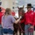 A U.S. Army soldier identified as SGT Hernandez from the Commanding General's Mounted Color Guard at Fort Riley, Kansas, is allowing a young woman to pet a dark brown horse at what appears to be a public outreach event. The soldier is wearing a distinctive red western shirt, black cavalry hat with gold insignia, and jeans. The event appears to be held near a stadium or university facility, likely Kansas State University based on the Wildcat logo visible on the building.