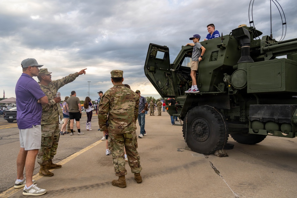 A military public display or open house event where civilians and uniformed soldiers interact around a large military vehicle, likely an M142 HIMARS or similar artillery rocket system. Two children have climbed on top of the olive-drab military vehicle while soldiers engage with civilian visitors on the ground. The event appears to take place on a paved lot or airfield under overcast skies.