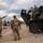 A military public display or open house event where civilians and uniformed soldiers interact around a large military vehicle, likely an M142 HIMARS or similar artillery rocket system. Two children have climbed on top of the olive-drab military vehicle while soldiers engage with civilian visitors on the ground. The event appears to take place on a paved lot or airfield under overcast skies.