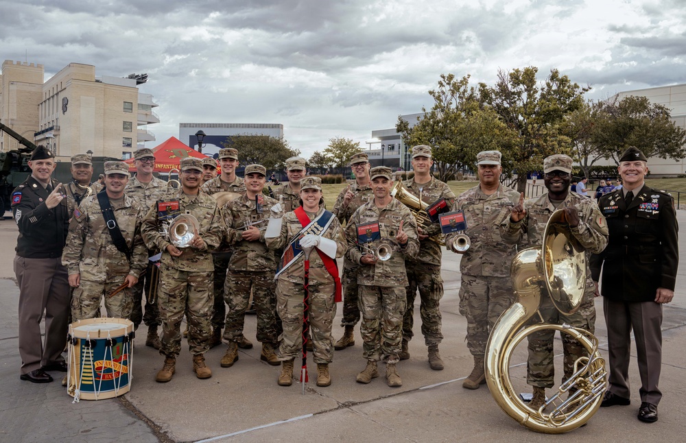 A military band group poses for a formal photograph outdoors on what appears to be a military installation or base. The personnel are holding various musical instruments including a sousaphone, drum, trumpets, and a baton, with one individual wearing a drum major sash. Two individuals on the far left and right appear to be in dress uniforms while the majority wear combat/utility uniforms (ACUs/OCP pattern).