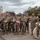 A military band group poses for a formal photograph outdoors on what appears to be a military installation or base. The personnel are holding various musical instruments including a sousaphone, drum, trumpets, and a baton, with one individual wearing a drum major sash. Two individuals on the far left and right appear to be in dress uniforms while the majority wear combat/utility uniforms (ACUs/OCP pattern).